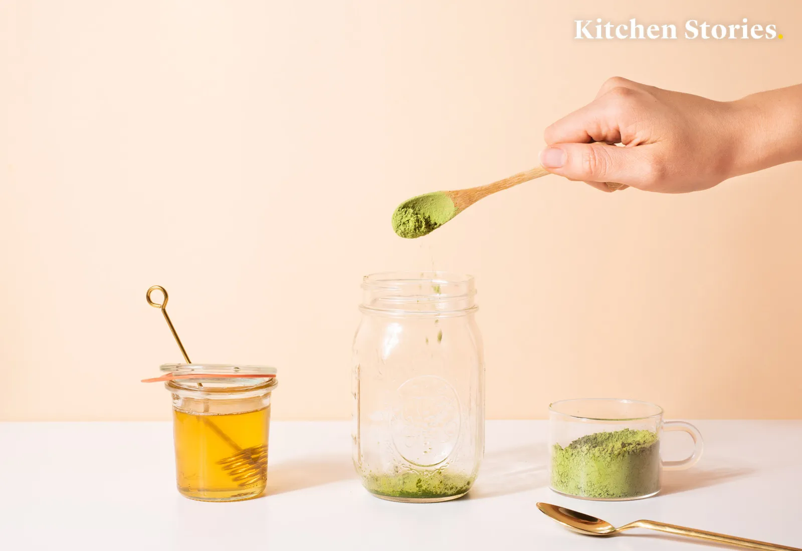 Hand adding matcha powder to a glass jar, with honey jar and spoon nearby on a white surface.