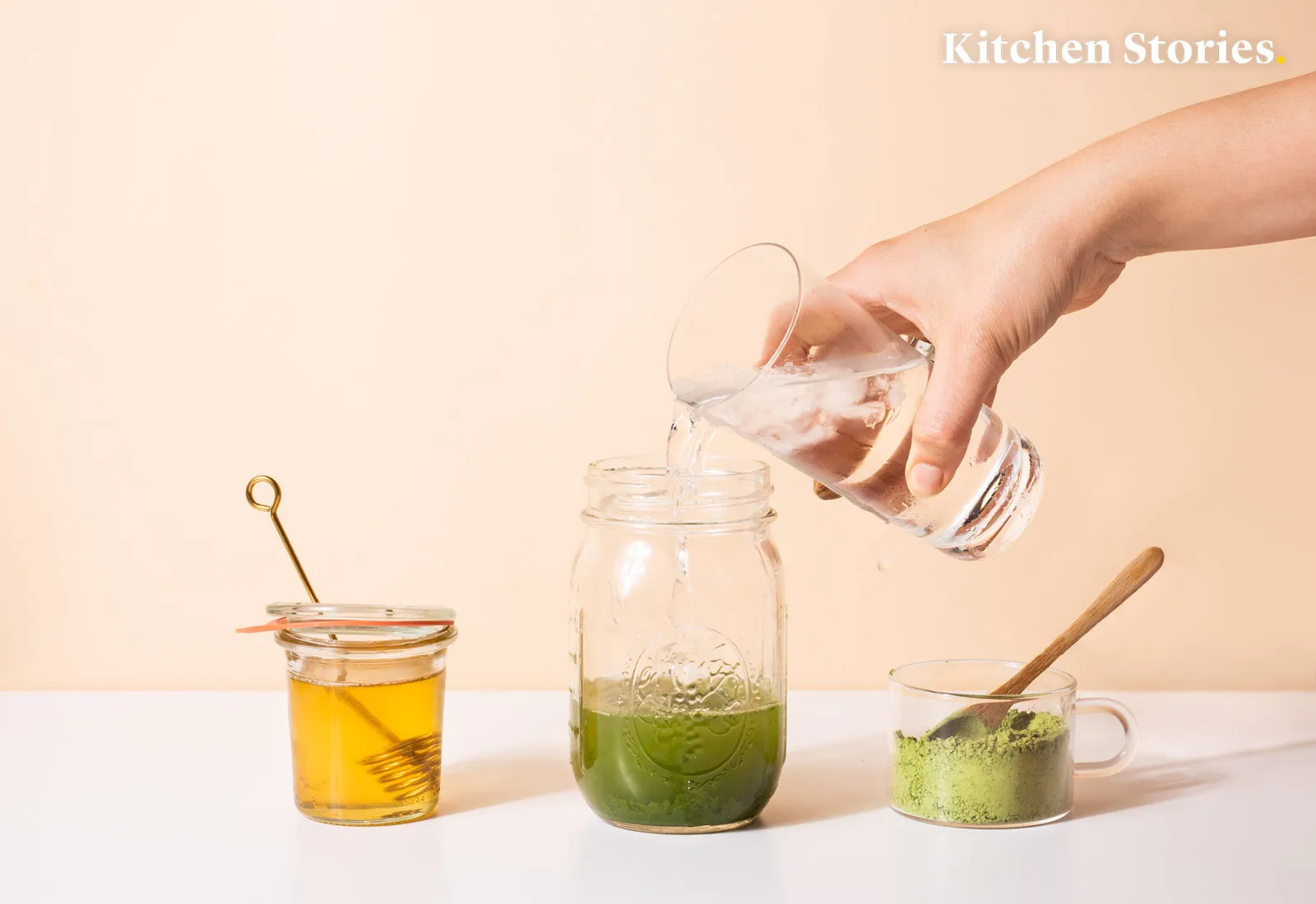 A hand pours water into a jar with green matcha, next to jars of honey and more matcha.