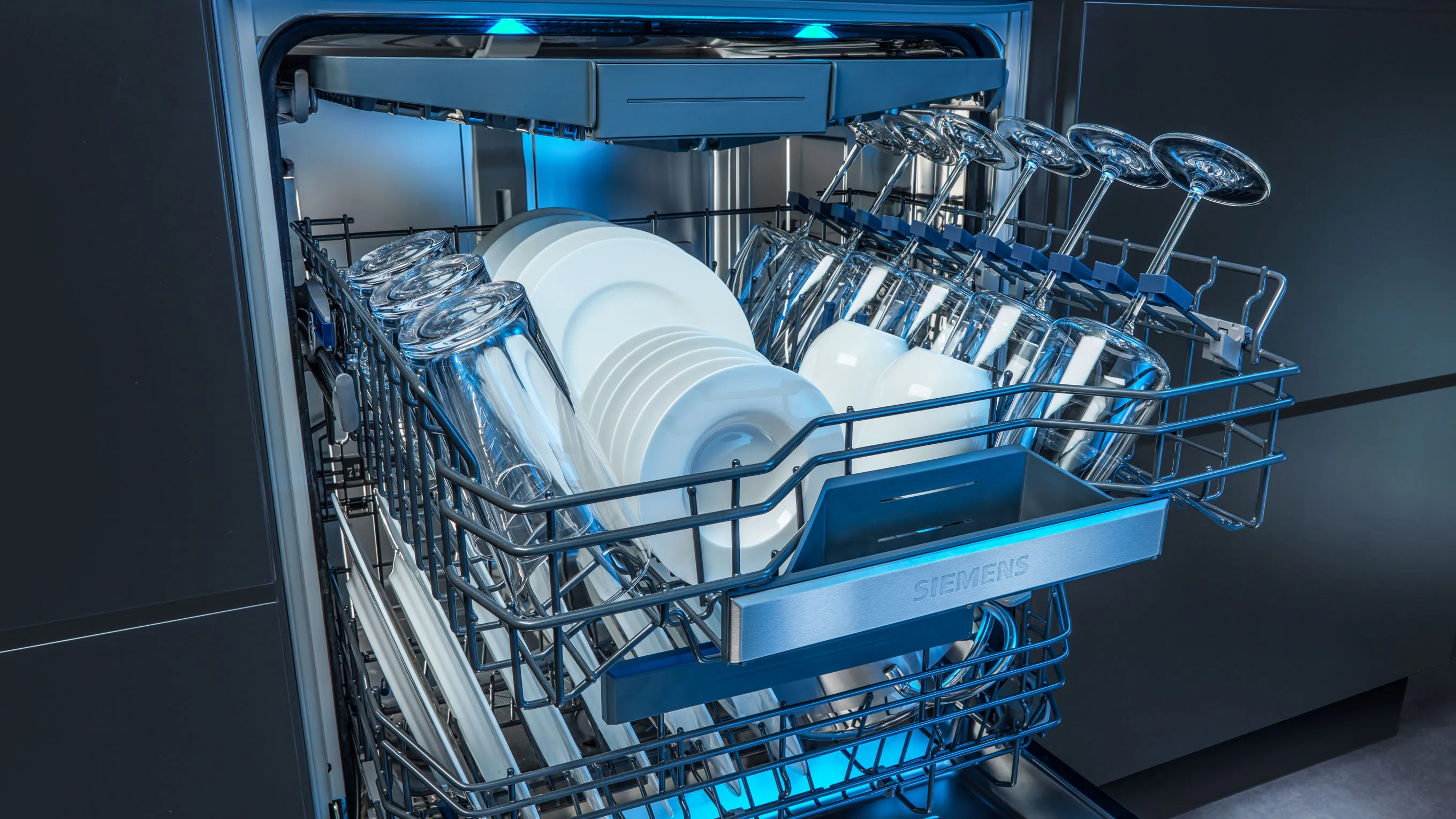 Dishwasher interior with clean dishes, plates, and utensils arranged in racks, illuminated by blue lighting.