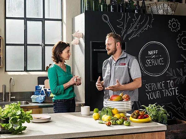 A NEFF technician in a gray polo shirt stands in a modern kitchen, interacting with a tablet near a sleek cooktop and bowls of fresh fruit.