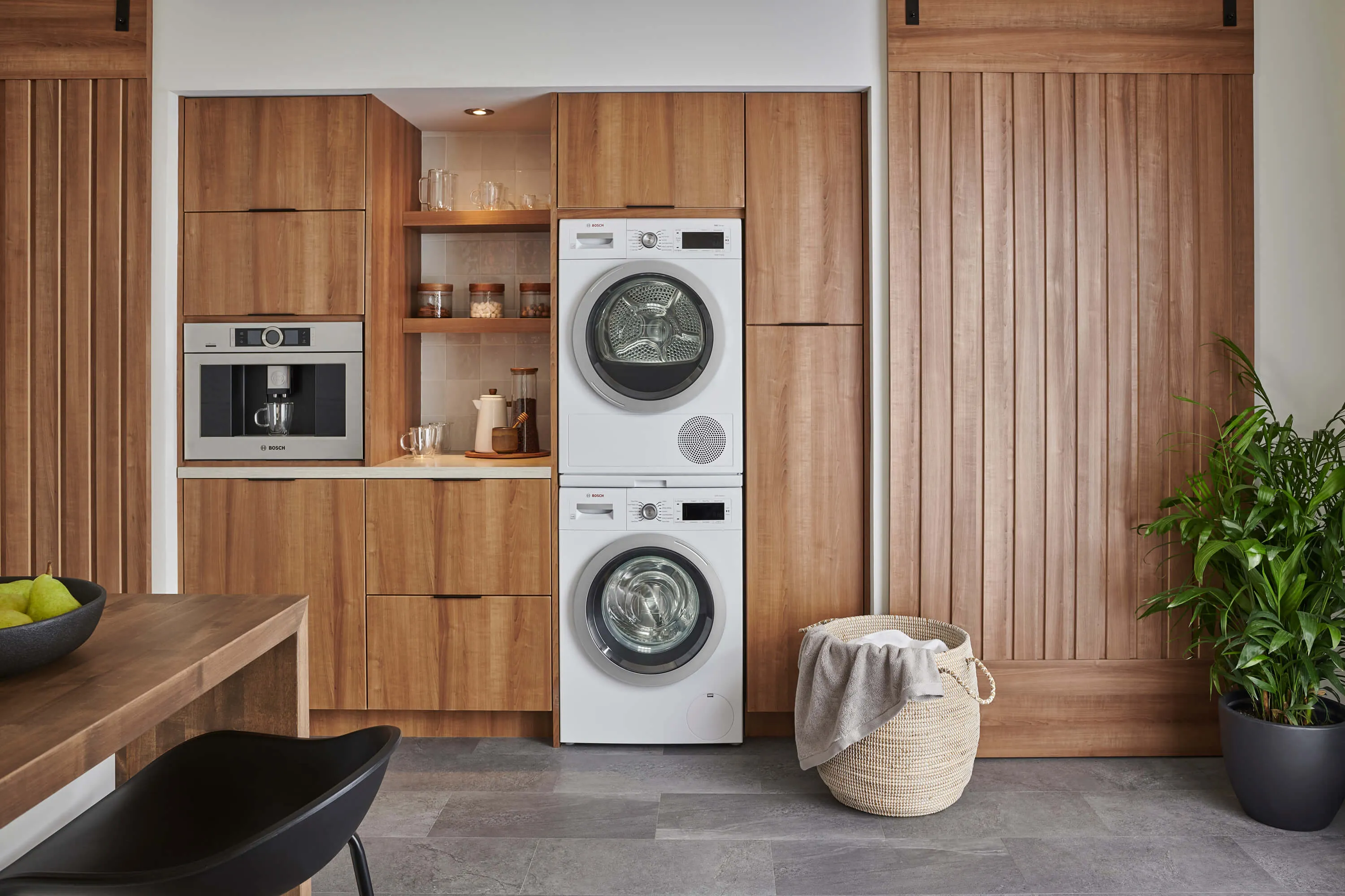 A modern laundry area with stacked Bosch washing machine and dryer units, integrated into warm wooden cabinetry with a built-in coffee machine and a laundry basket.