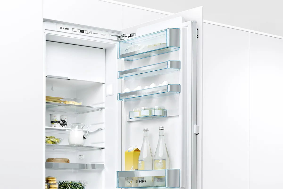 A modern white refrigerator stands open in a bright kitchen. Inside are mostly empty glass shelves with a few food items, a white teapot, and containers. The door shelves hold two glass bottles, a carton of orange juice, and a few small jars.