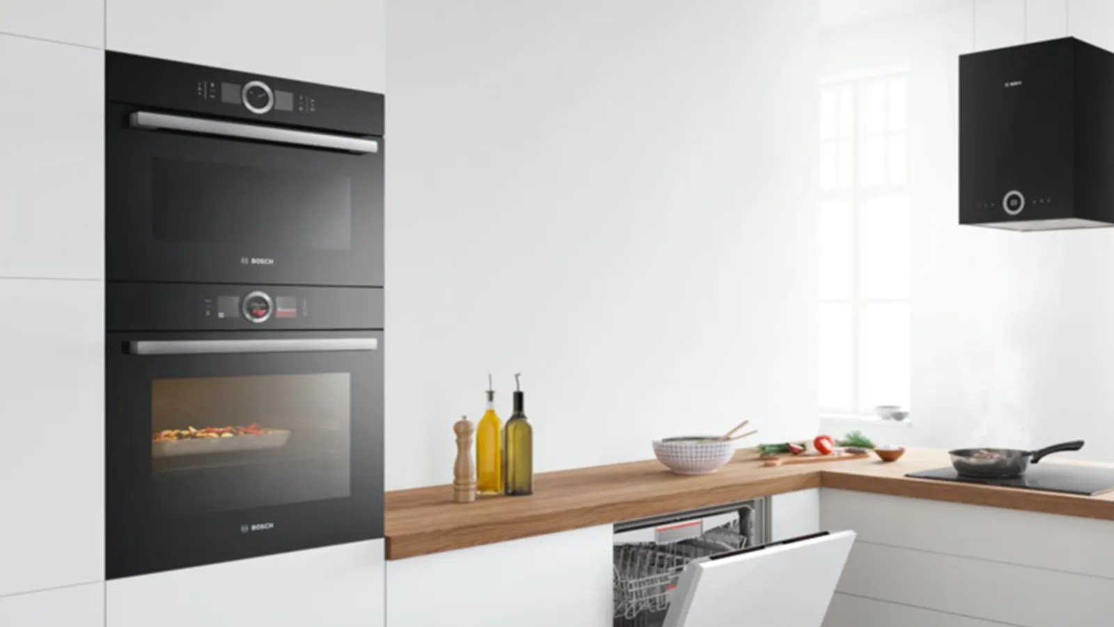 A modern kitchen with integrated black Bosch appliances, including a stacked oven and microwave, an open dishwasher, and a black extractor hood, all set against white walls and light wooden countertops.