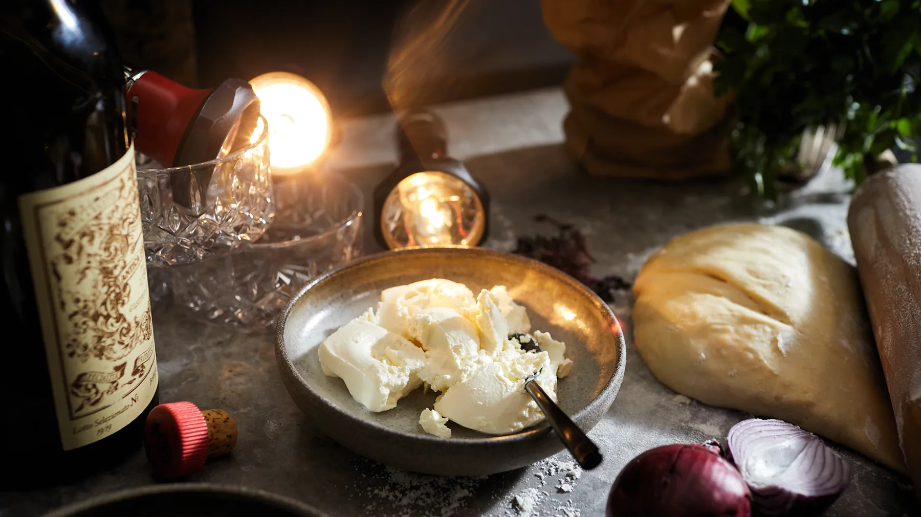 Рустик маса с вино, хляб, сирене и други хранителни продукти в топло осветление.