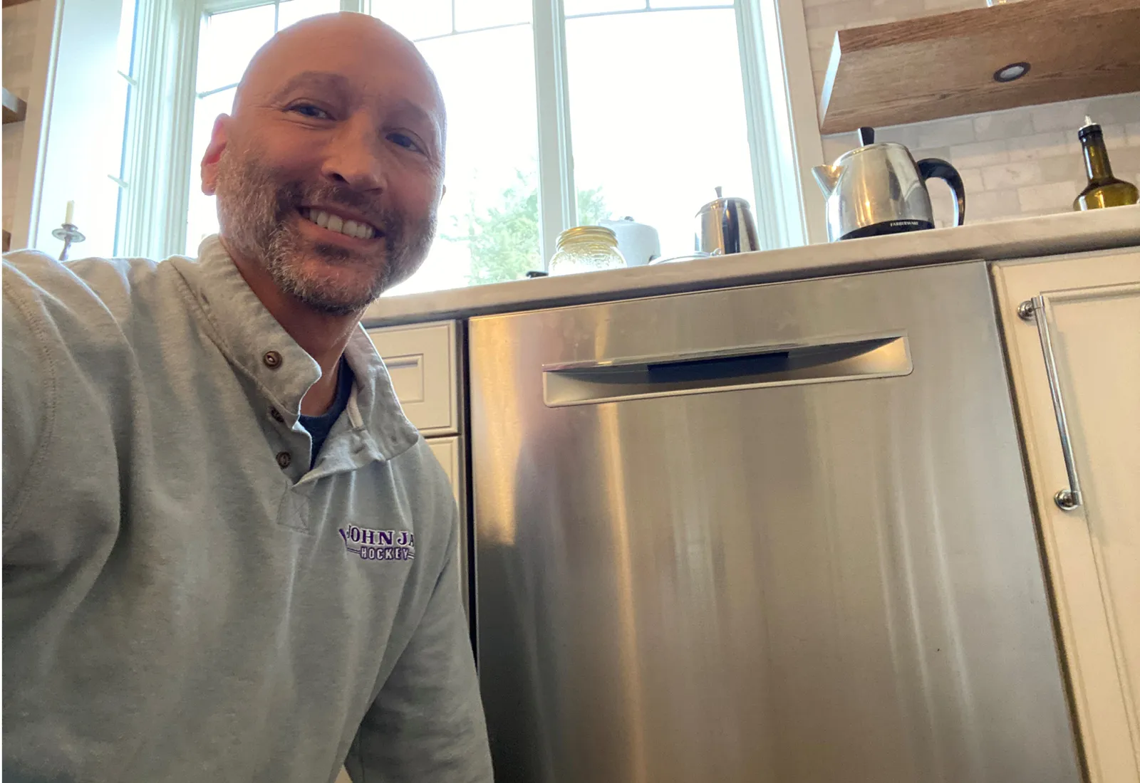 Daniel smiling next to a stainless steel dishwasher in a bright kitchen with large windows.