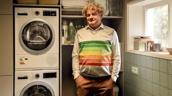Un hombre sonriente de pelo rizado, con un suéter de rayas de colores, está de pie junto a una pila de lavadoras en un cuarto de servicio luminoso y ordenado.