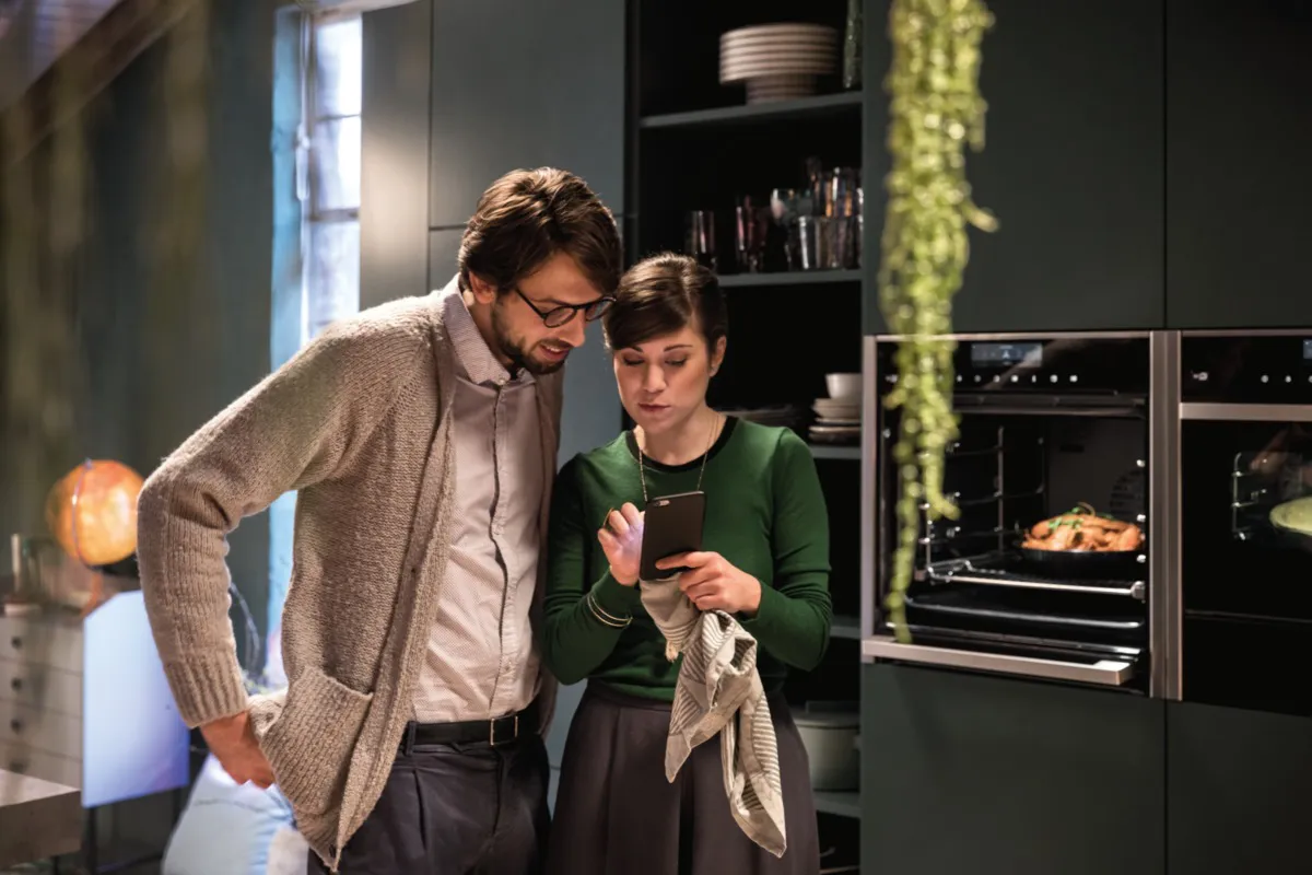 Two people stand in a modern kitchen, leaning over a smartphone; the woman holds a dish towel while a roast cooks in the oven behind them.