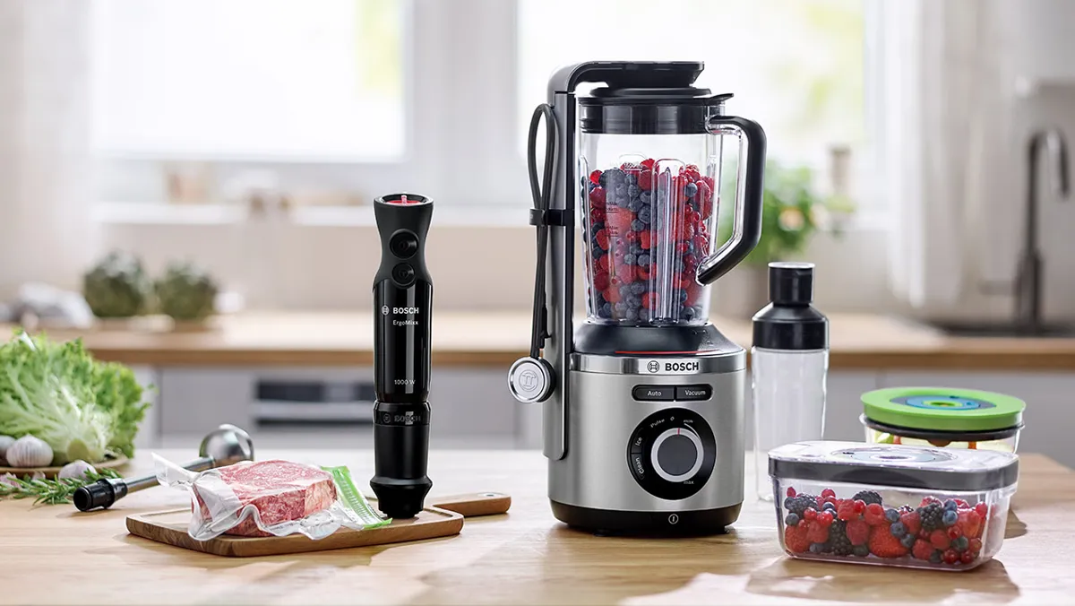 Küchengeräte einschließlich Mixer, Lebensmittelverarbeiter und Aufbewahrungsbehälter auf Holztisch mit frischen Produkten.