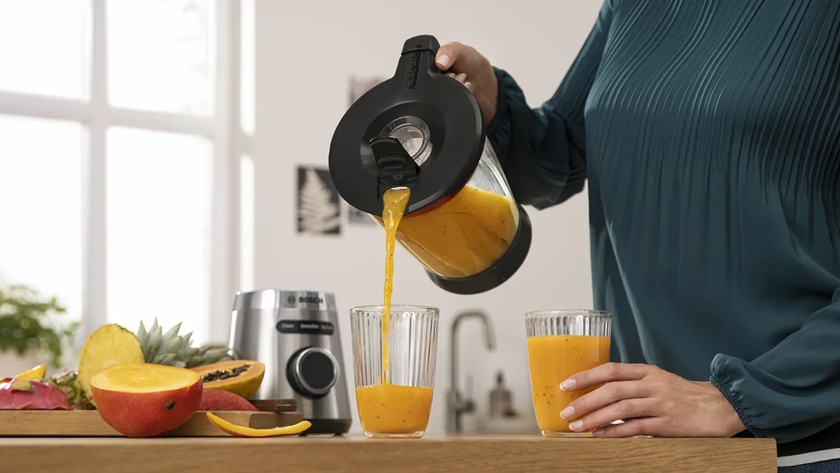 Persona che versa del succo d'arancia fresco nei bicchieri sul tavolo della cucina insieme alla frutta e al frullatore.