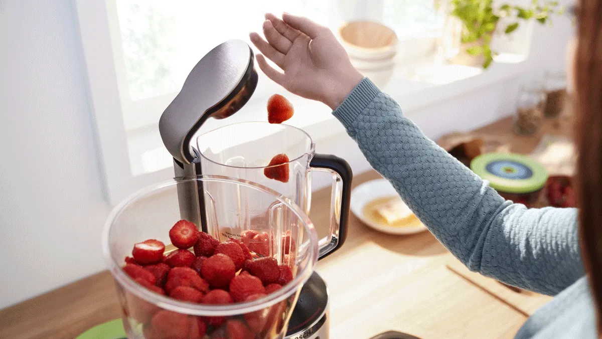 Miscelatore da cucina con verdure fresche, donna in sottofondo che prepara il cibo.