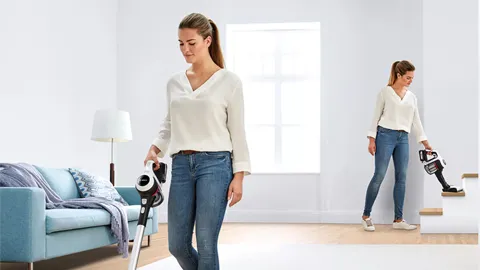 Woman in white sweater vacuuming living room, another woman in background.