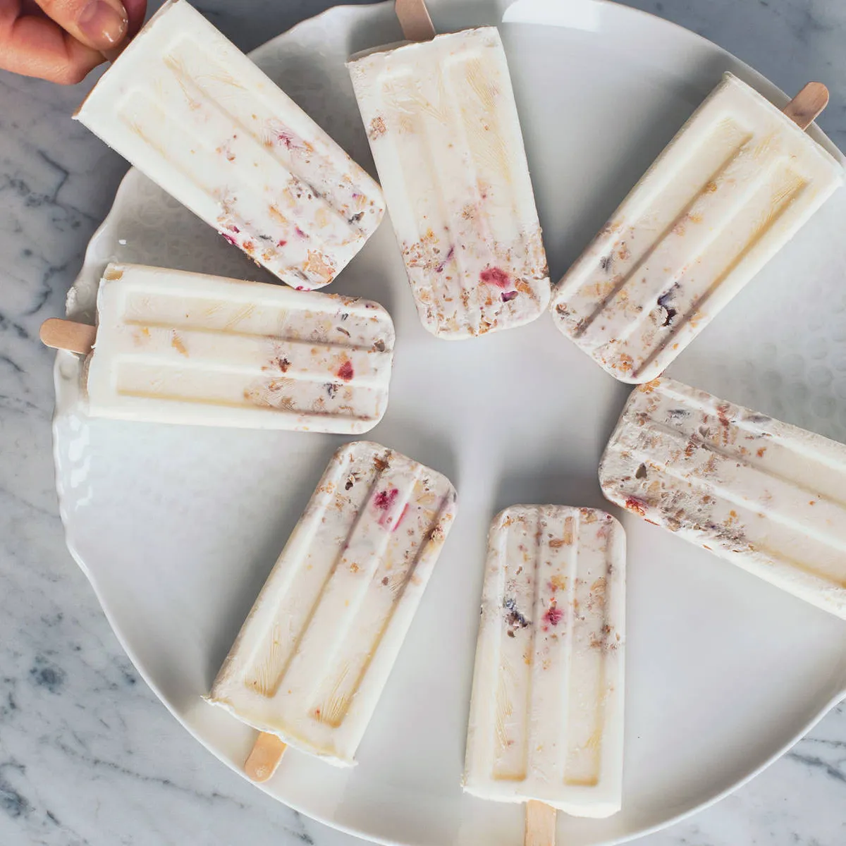 Seven creamy white popsicles with visible fruit pieces and granola, arranged in a circle on a white round plate on a marble surface; a hand holds one at the top left.
