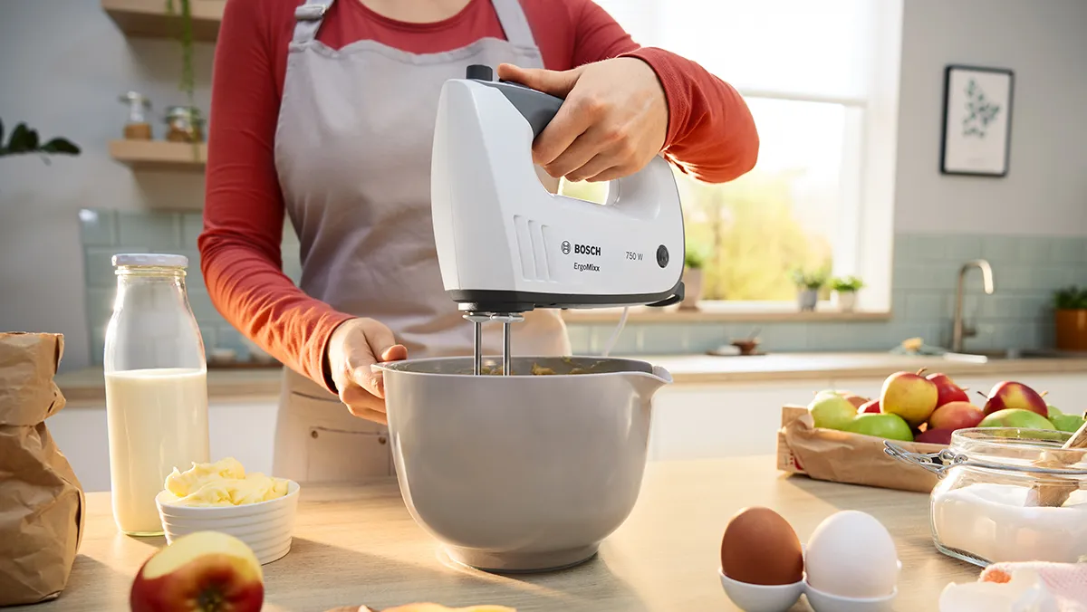 A person uses a Bosch hand mixer to beat the contents of a white bowl.