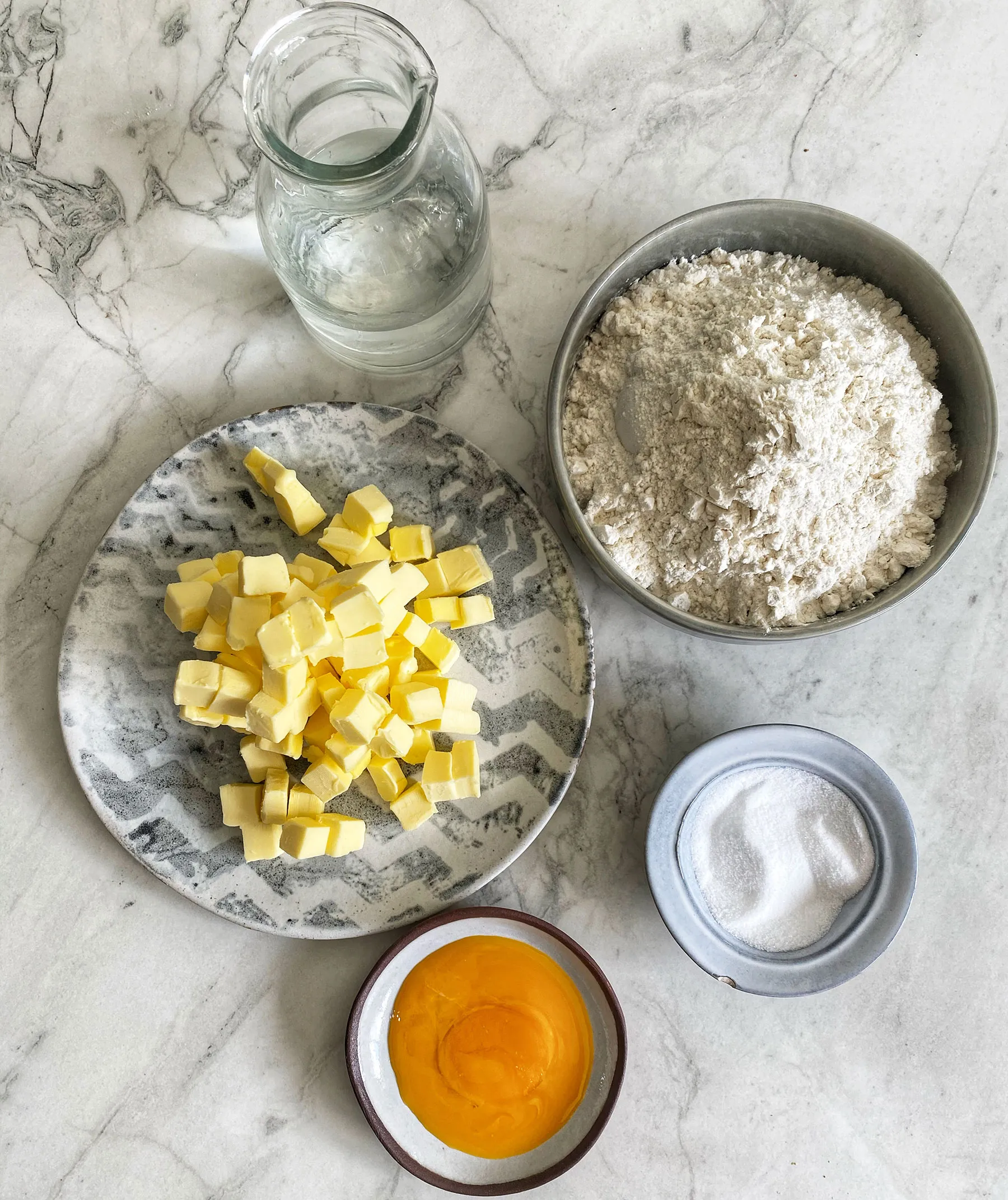 An overhead flat lay of ingredients for pastry: a plate of diced butter, a bowl of flour, a bowl of sugar, an egg yolk in a small bowl, and a jug of water, all on a white marble surface.