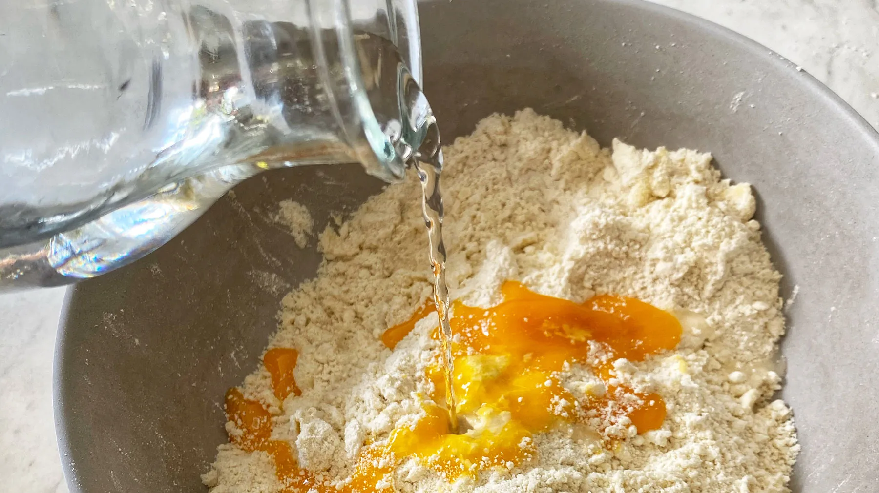 Water is poured from a glass jug into a grey bowl containing flour and egg yolks, preparing dough.