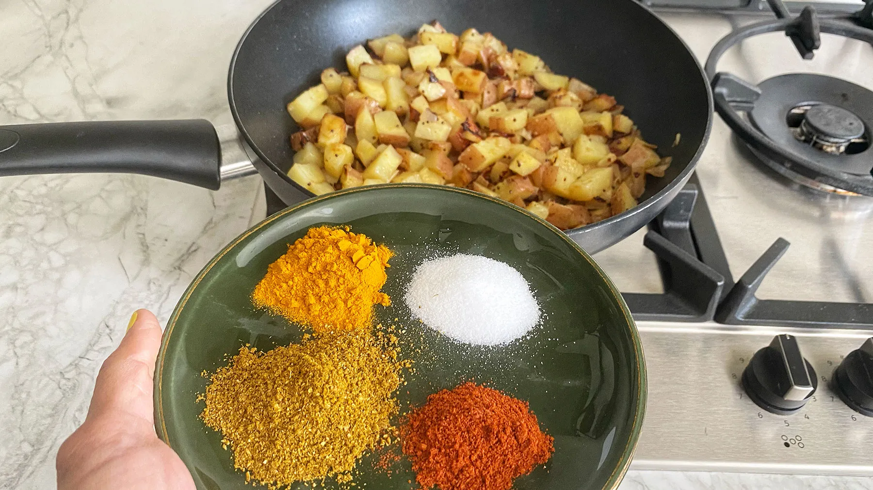 A hand holds a green plate with piles of turmeric, curry powder, chili powder, and salt, ready to be added to a pan of cooking potatoes on a gas hob.