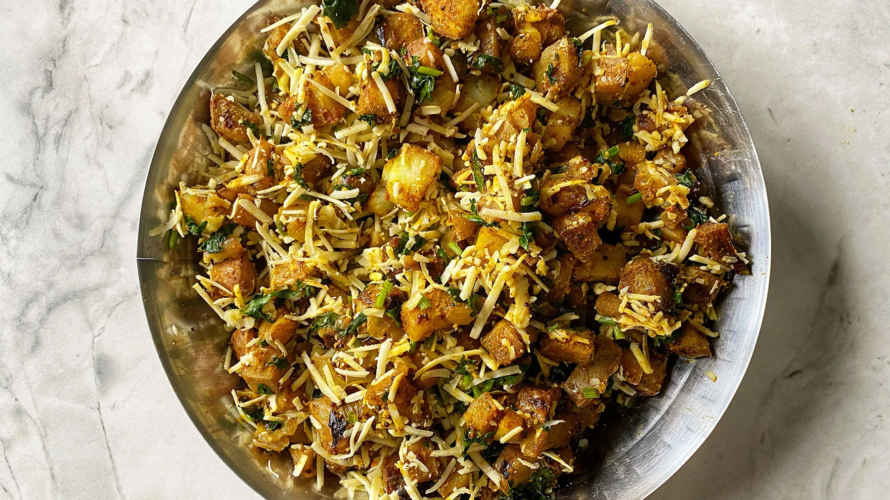 An overhead view of a metal bowl filled with diced roasted potatoes, shredded cheese, and chopped green herbs.