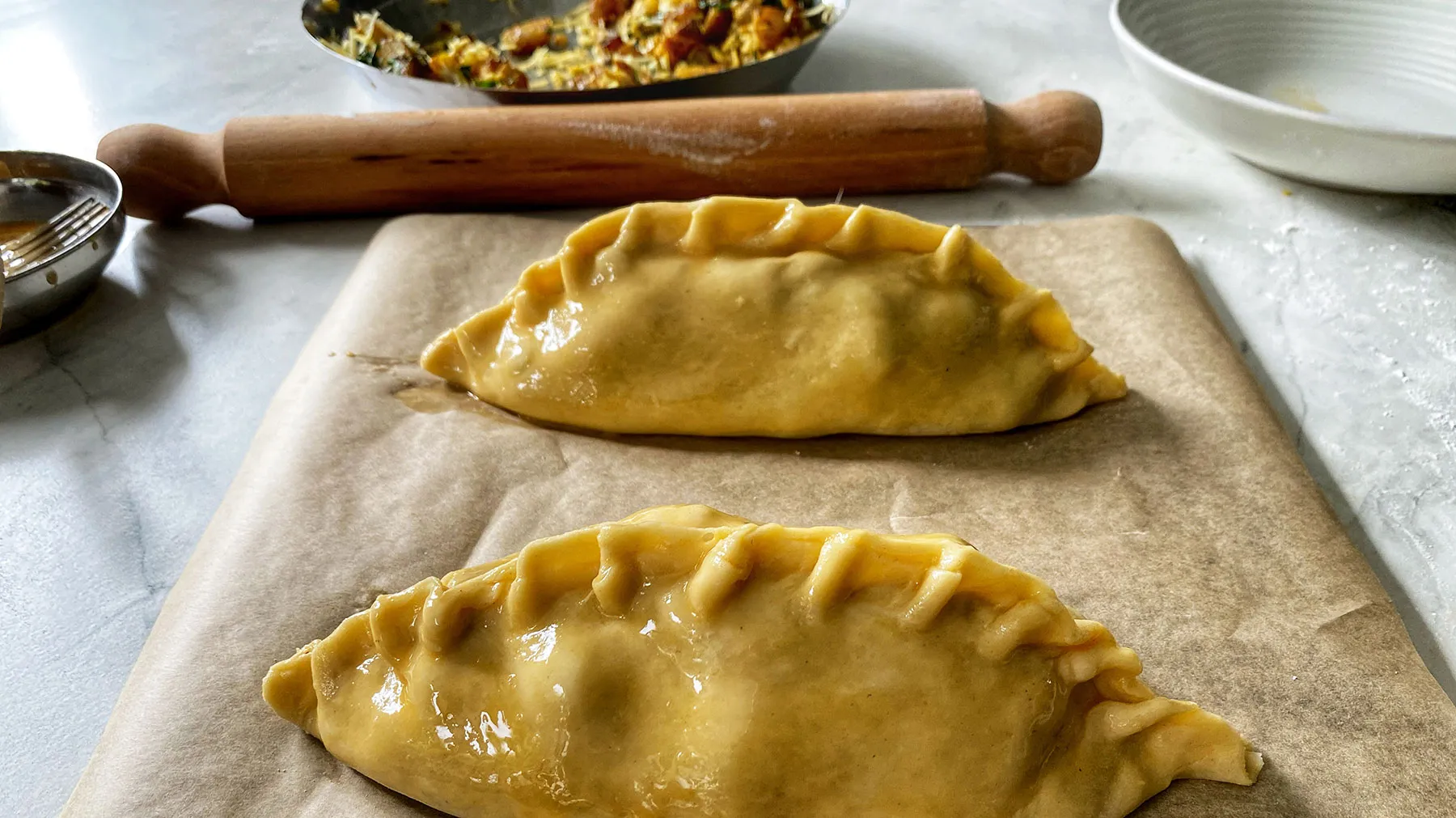 Two uncooked pasties with crimped edges, brushed with egg wash, resting on parchment paper on a baking sheet, with a rolling pin and filling in the background.