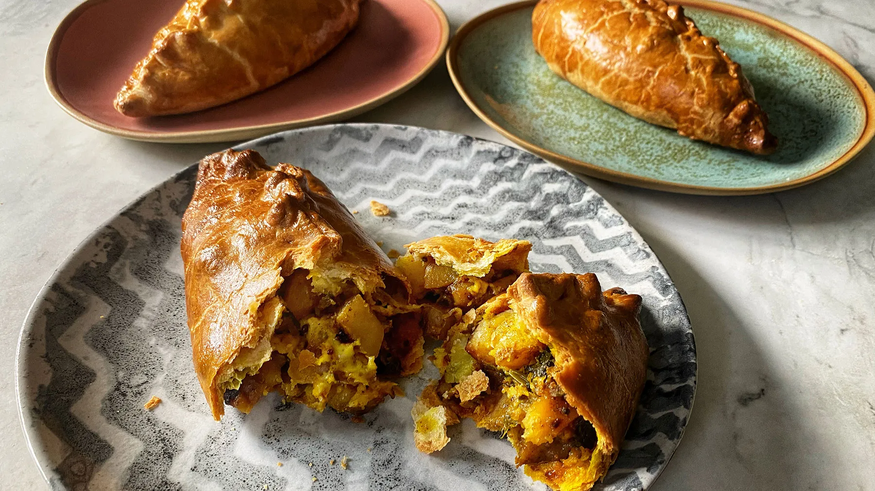Three golden-brown pasties on plates, one broken open to reveal a savory potato and vegetable filling, on a white marble surface.