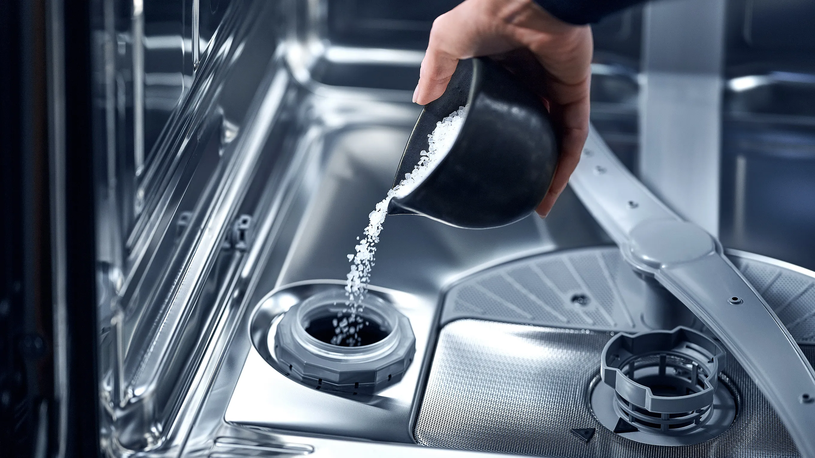 A hand pours white dishwasher salt from a small, black ceramic cup into the salt reservoir at the bottom of a stainless steel dishwasher.