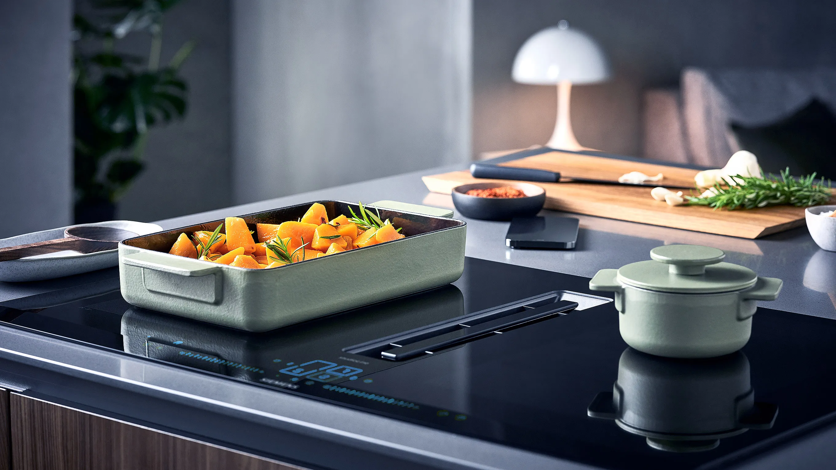 Modern kitchen stovetop with two mint-green pots, one filled with roasted vegetables and herbs, and a cutting board with spices and rosemary.