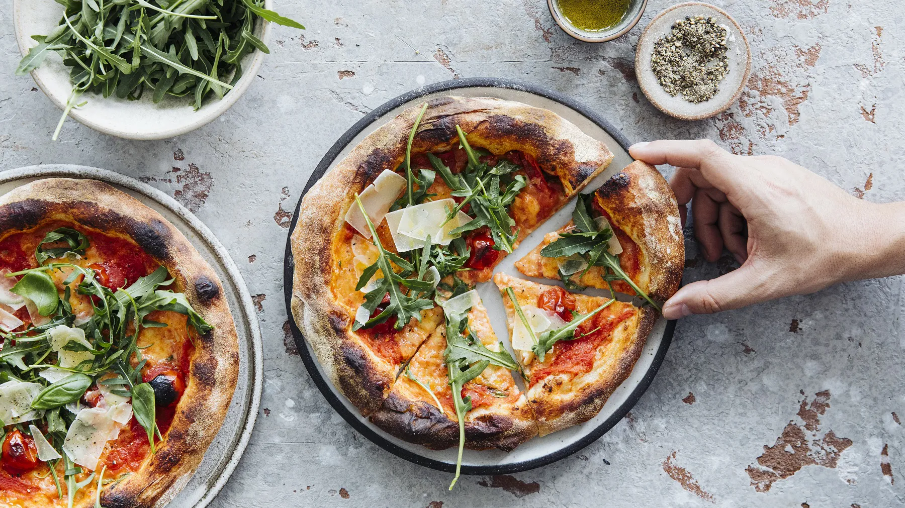 Nahaufnahme einer neapolitanischen Pizza mit Rucola und Parmesanspänen auf einem dunklen Teller, daneben eine Hand, die ein Stück hält