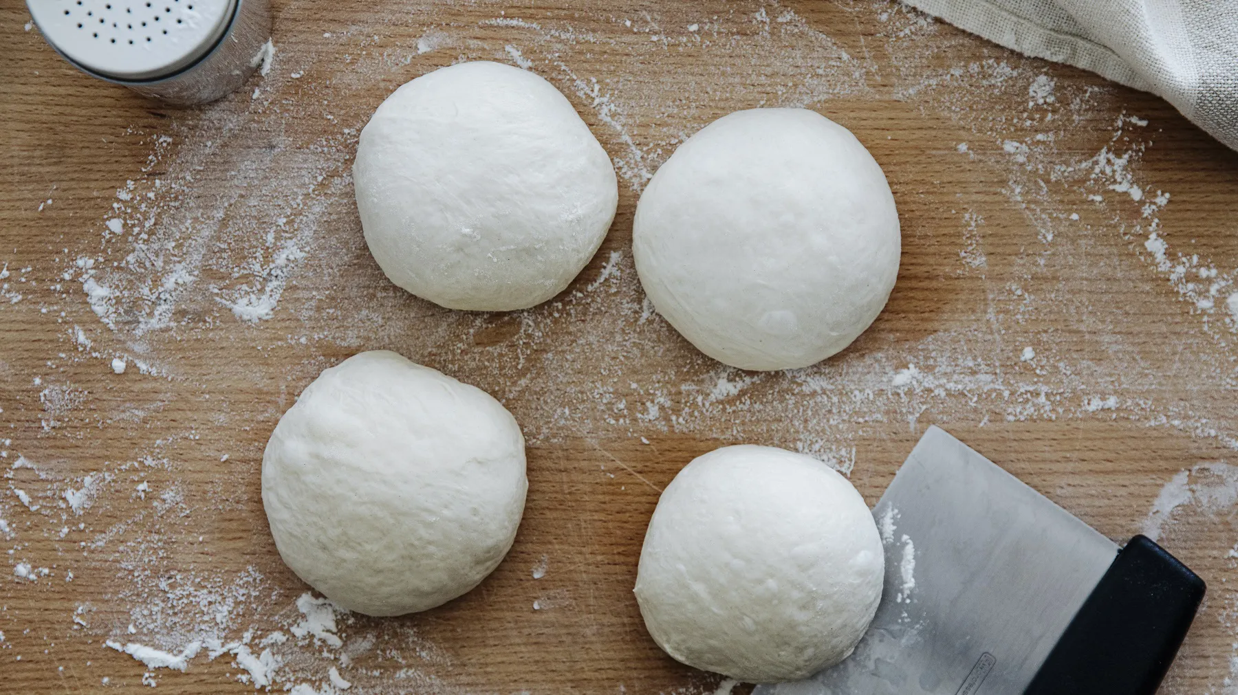 Vier runde Pizzateigkugeln auf bemehltem Holzbrett mit Teigschaber