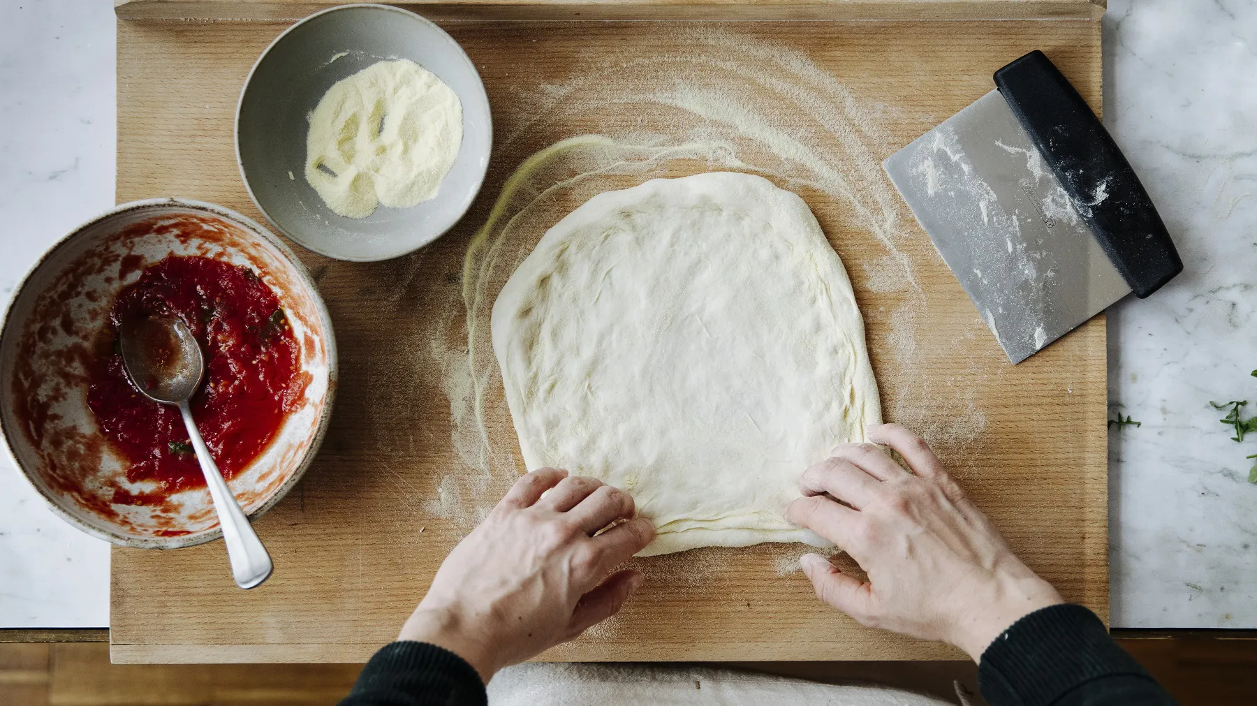 Hände formen einen rechteckigen Pizzateig auf einem Holzbrett neben einer Schüssel mit Tomatensauce, einer Schüssel mit Mozzarella und einem Teigschaber