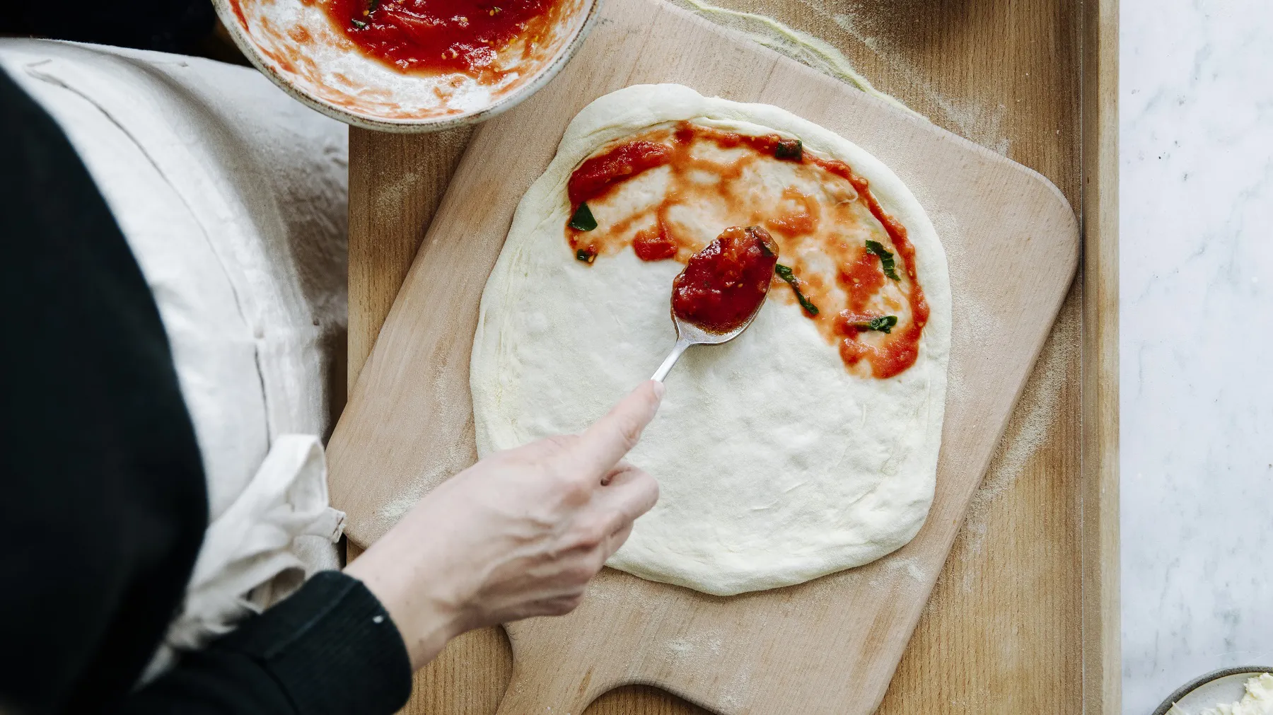 Hand streicht Tomatensauce mit Löffel auf runden Pizzateig auf Holzbrett, Schüssel mit Tomatensauce daneben