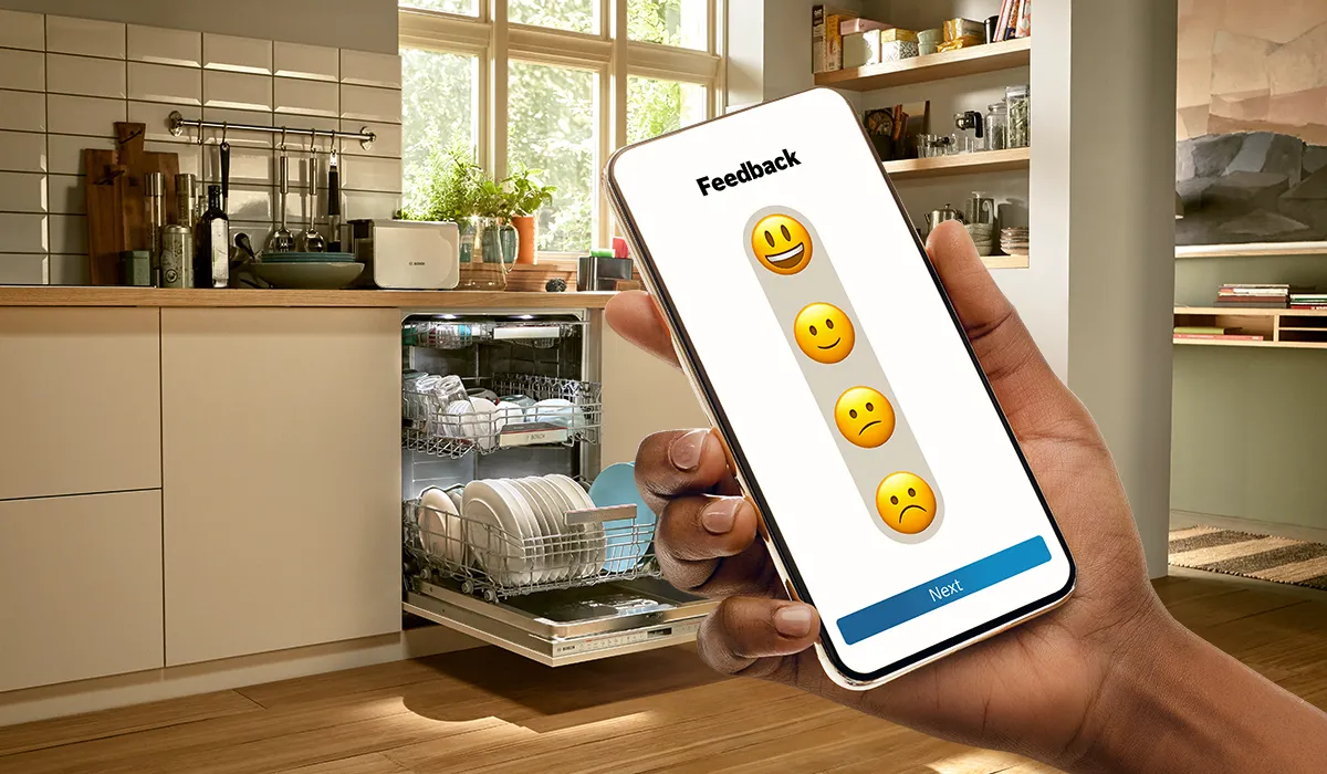 Ruka koja drži pametni telefon koji prikazuje aplikaciju za povratne informacije s emotikonima, perilica posuđa u pozadini kuhinje.