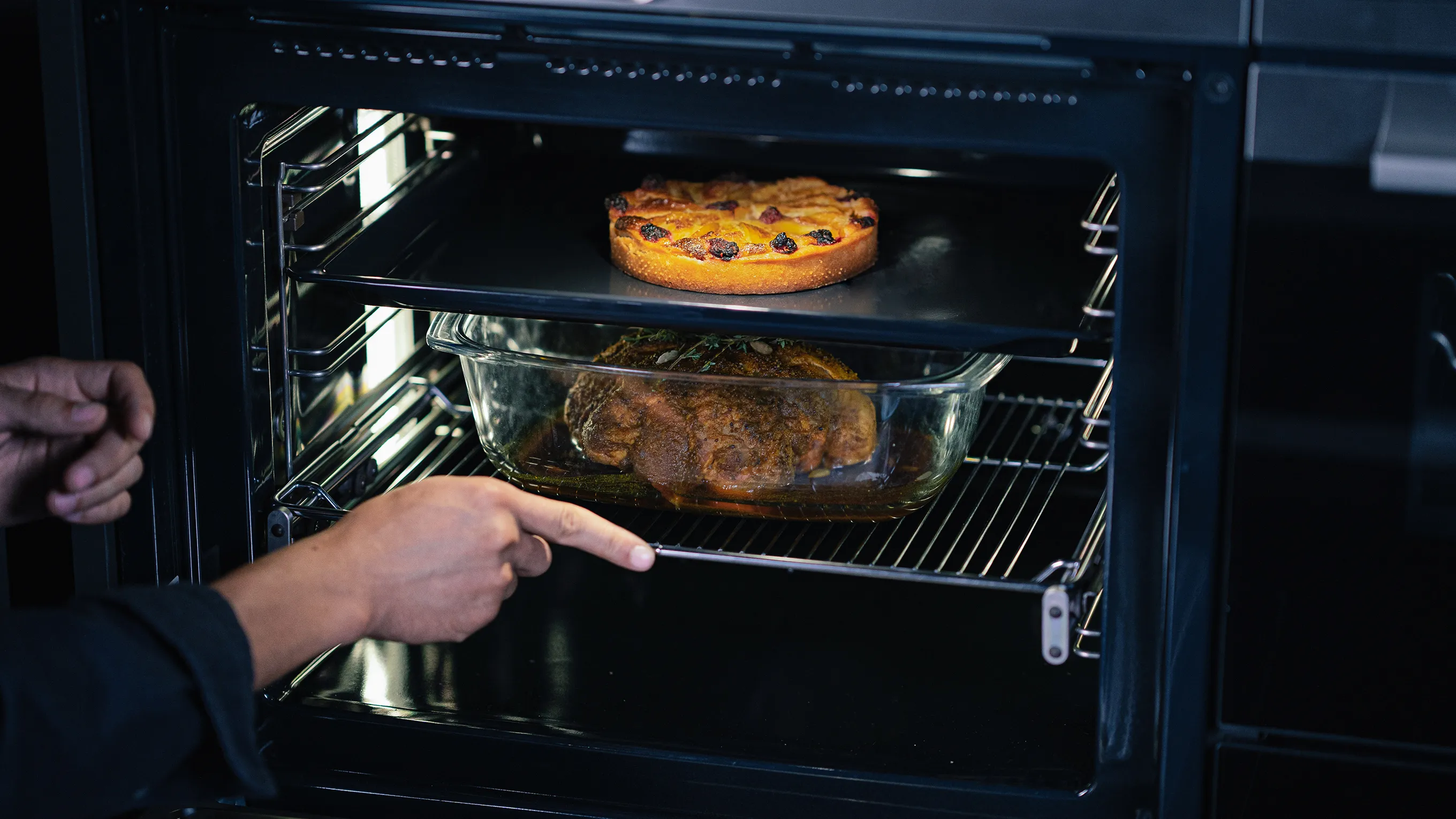 Oven interior with baking pizza and meat dish, hand reaching to check on food.