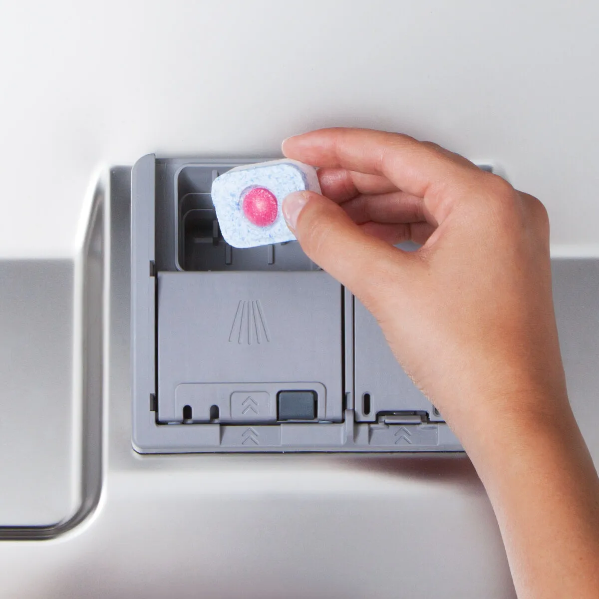 Hand placing a dishwasher tablet into a detergent compartment on a stainless steel dishwasher.