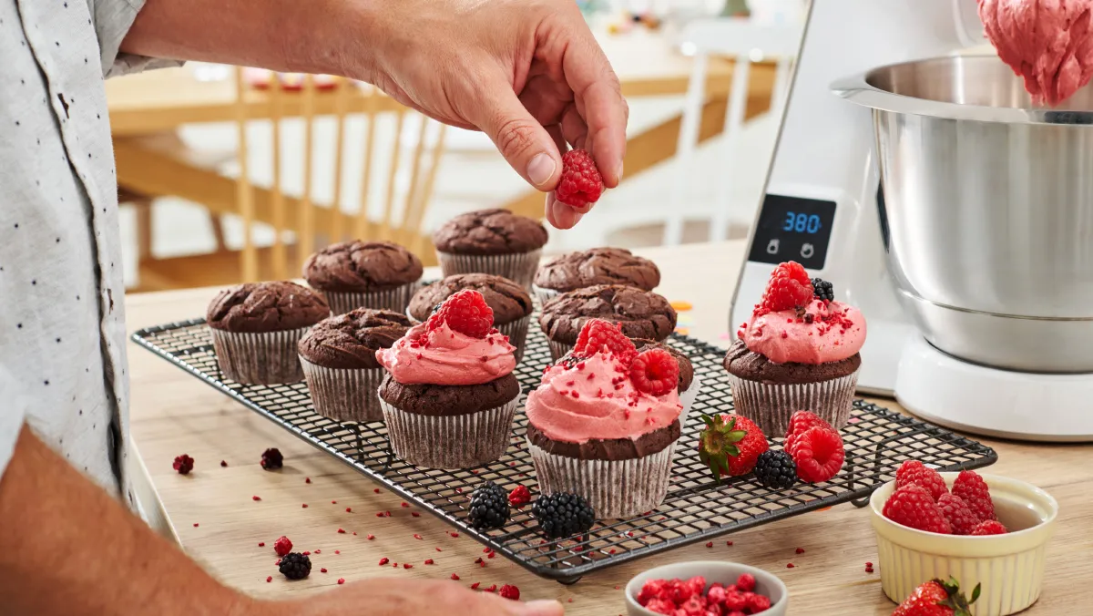 A hand places a fresh raspberry on a chocolate cupcake topped with pink frosting, surrounded by other cupcakes and berries on a cooling rack.