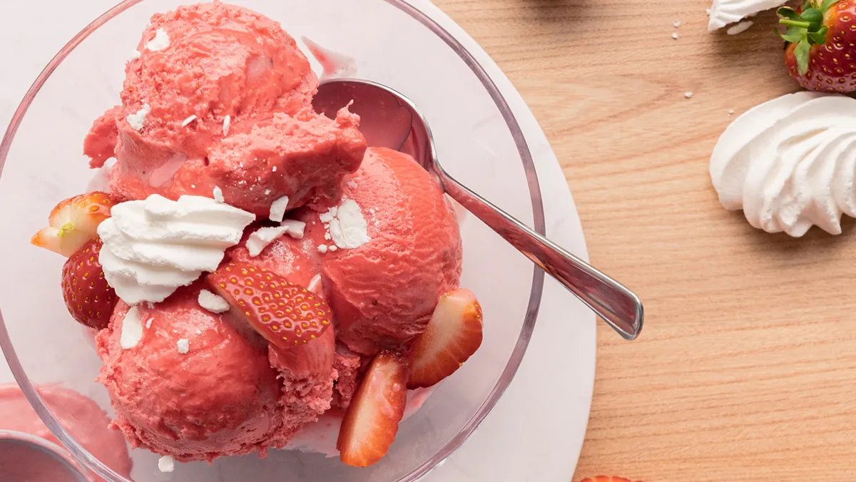 An overhead view of a glass bowl filled with scoops of pink strawberry ice cream, fresh strawberry slices, and white meringue pieces, with a spoon.
