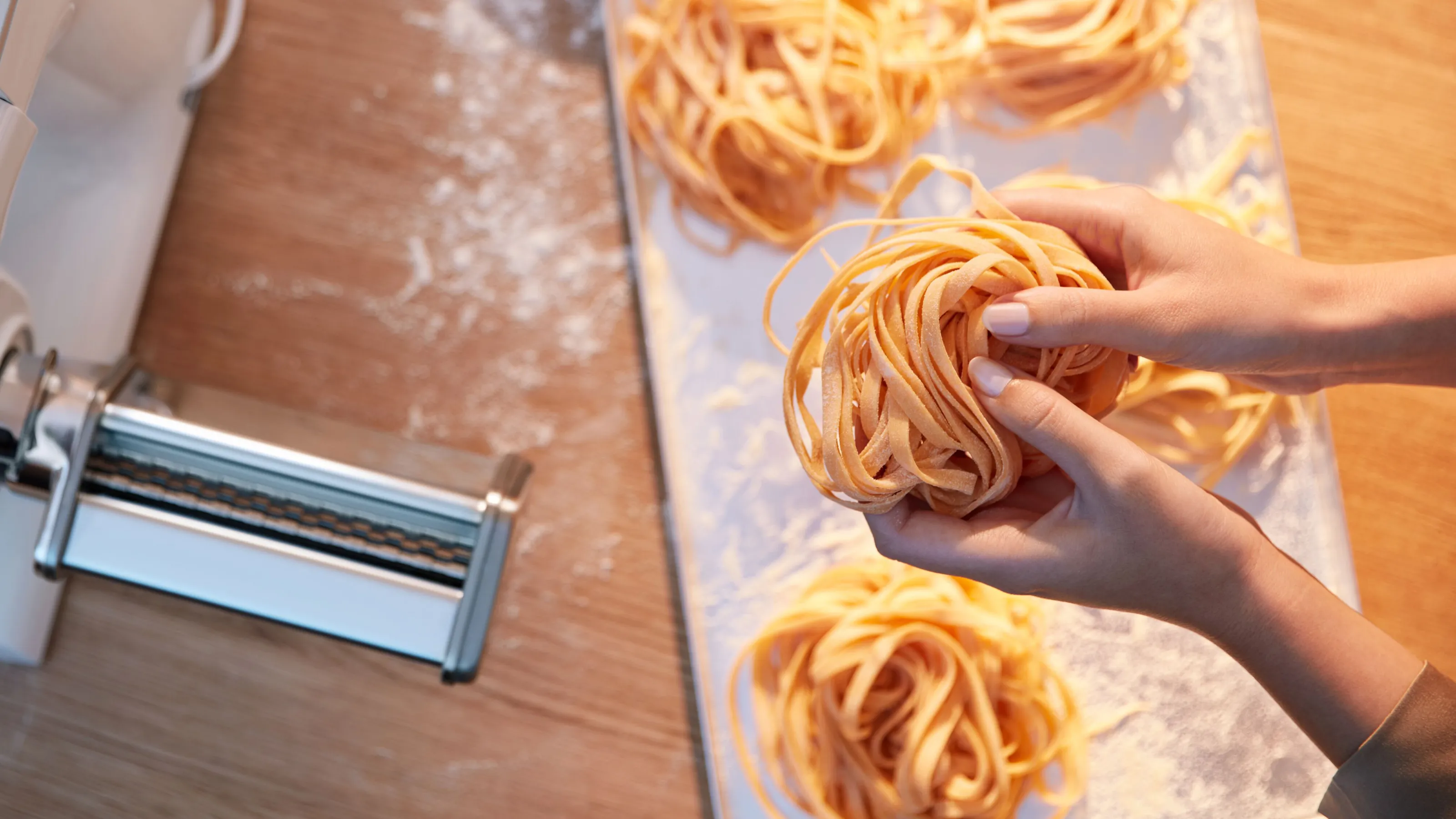 Hands gently shape fresh, homemade tagliatelle pasta into nests on a floured white board, next to a pasta maker attachment on a stand mixer.