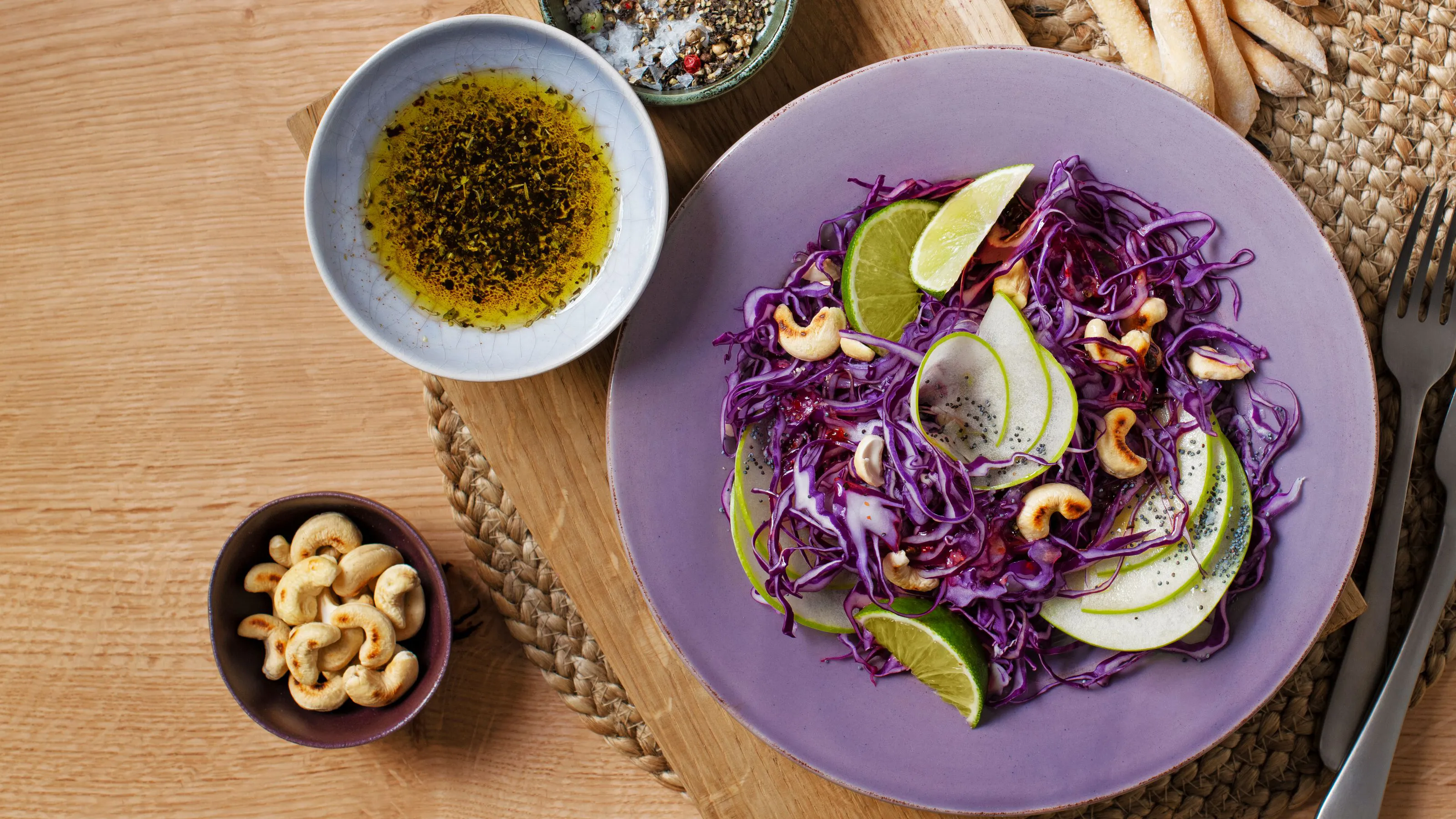 An overhead view of a vibrant red cabbage salad with green apple slices, cashews, and lime wedges on a purple plate, next to a bowl of dressing and roasted cashews.