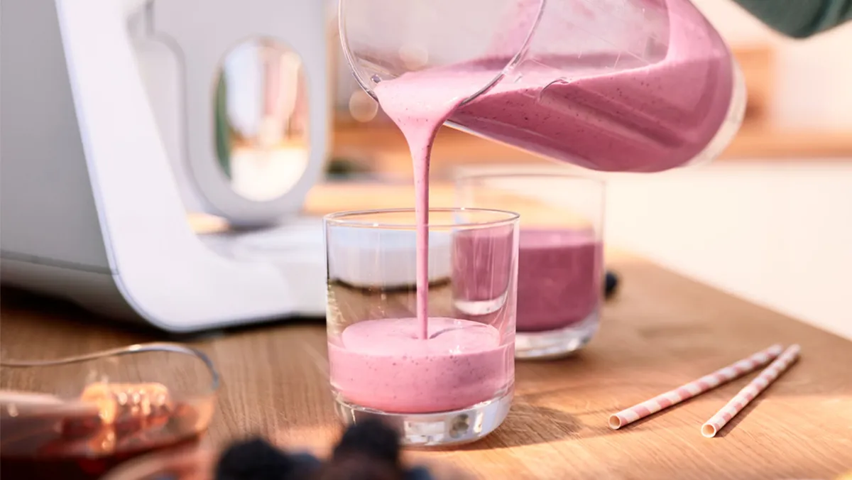 Pouring pink smoothie from a pitcher into glasses on a wooden table, with straws and a blender in the background.