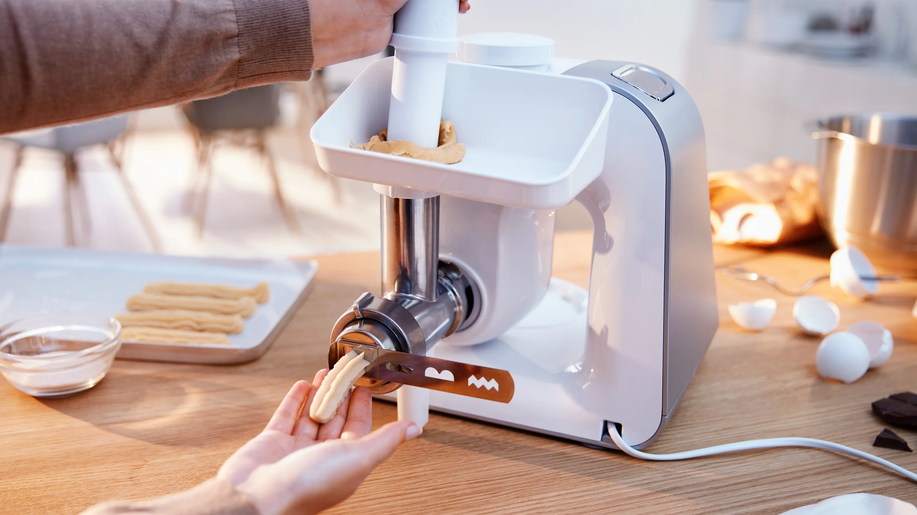 Hands operate a white Bosch stand mixer with a meat grinder attachment, extruding dough into churro shapes onto a baking tray.