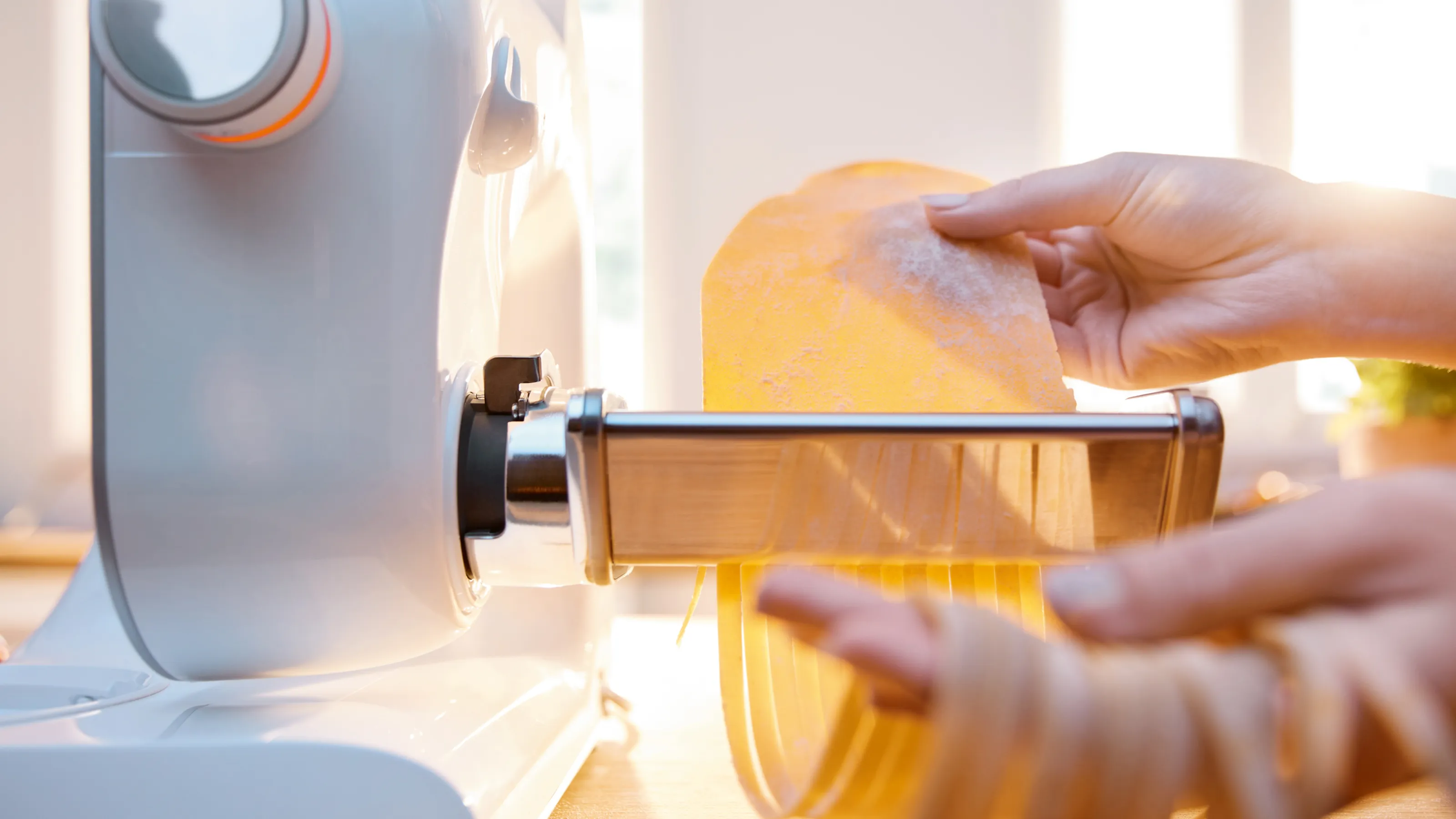 Hands feed a sheet of yellow pasta dough into a pasta maker attachment on a white stand mixer, producing fresh spaghetti.