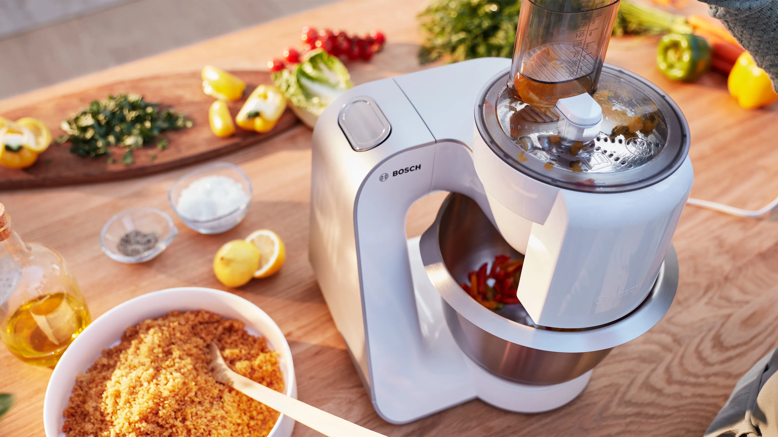 A white Bosch stand mixer with a food processor attachment is actively chopping red chili peppers, surrounded by fresh ingredients like herbs, lemons, and couscous on a wooden countertop.