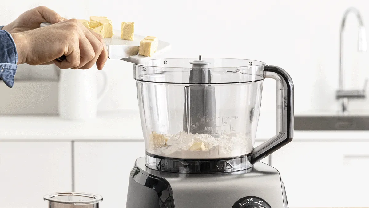 Una persona che aggiunge pezzi di burro a un robot da cucina pieno di farina, preparandosi a mescolare gli ingredienti in una cucina moderna.