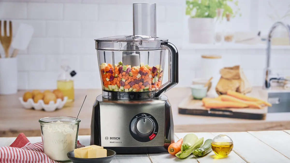 Un robot da cucina Bosch con verdure miste tritate sul bancone, circondato da ingredienti come formaggio, erbe e olio d'oliva.