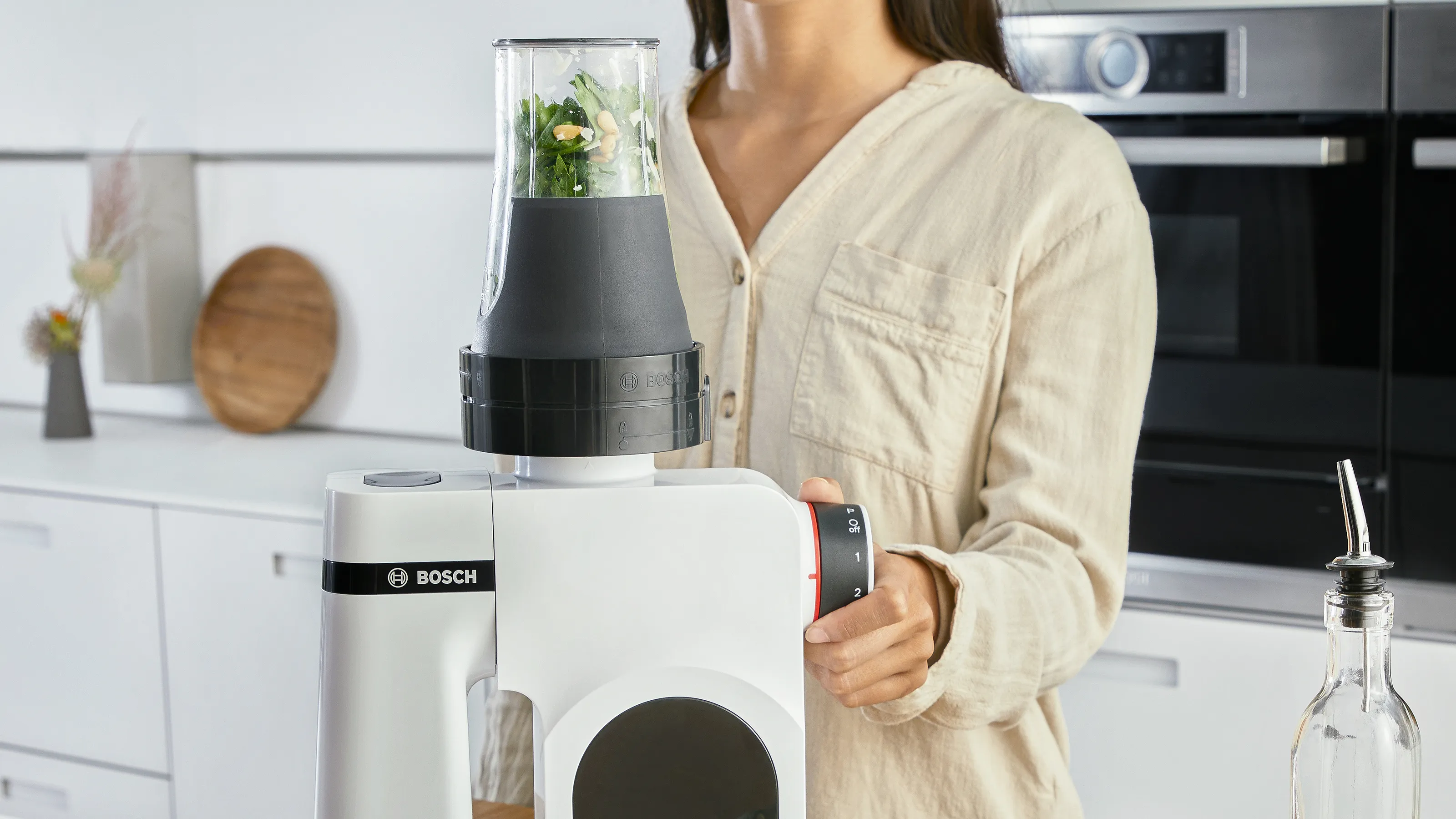A woman operates a white Bosch stand mixer with a blender attachment filled with green herbs and nuts, in a modern kitchen.