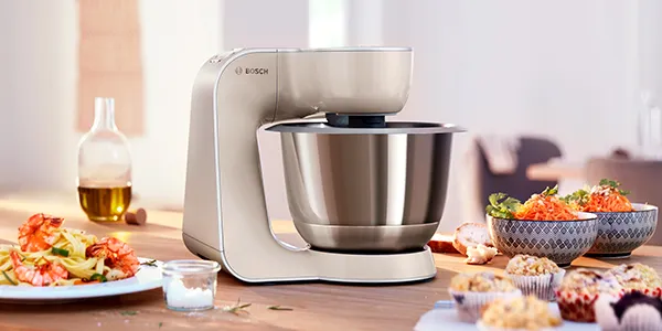 Un mixer da cucina Bosch con una ciotola in acciaio inossidabile si trova su un tavolo di legno, circondato da vari piatti pronti.
