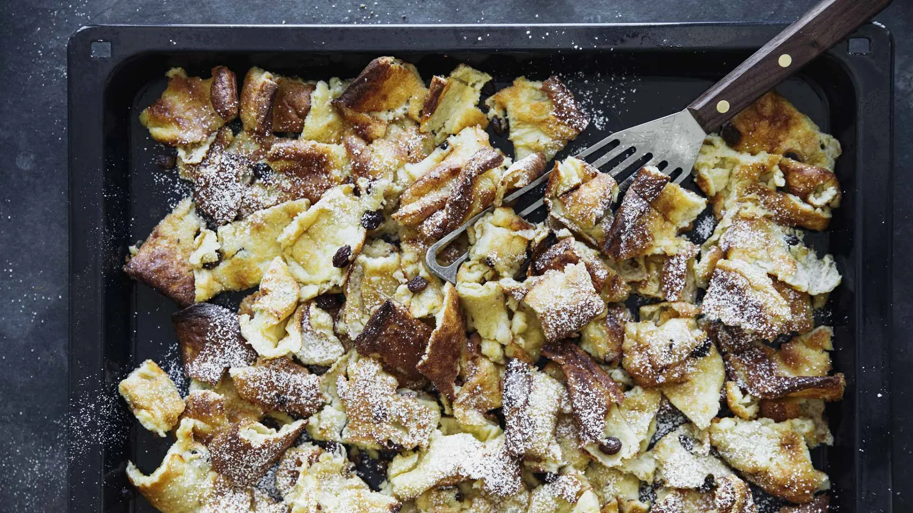 Backblech mit zerzupftem Kaiserschmarrn, bestreut mit Puderzucker und einer Metallschaufel darin