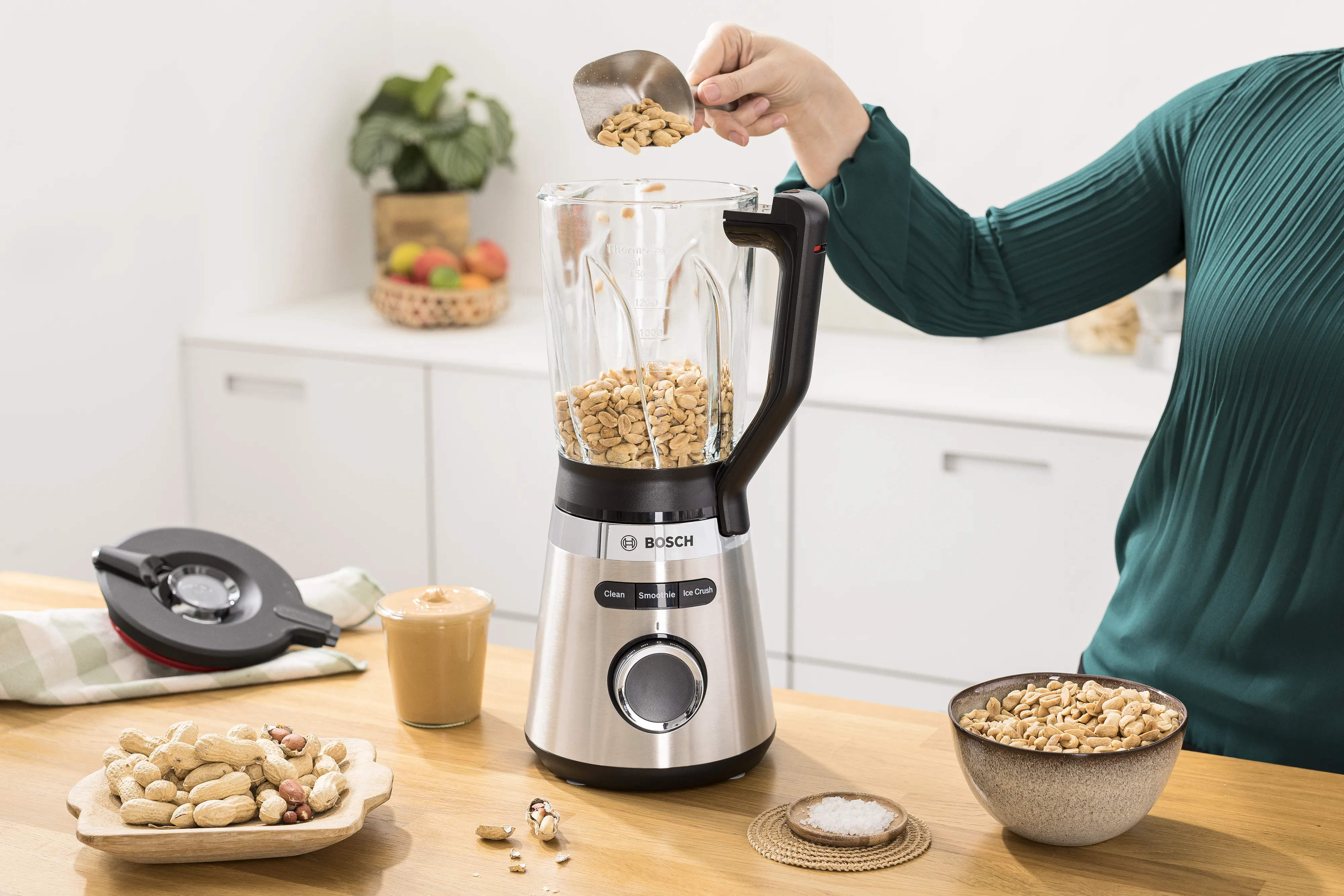 O persoană care pune alune într-un blender așezat pe blatul de bucătărie, având lângă ea boluri cu alune și un pahar cu unt de alune