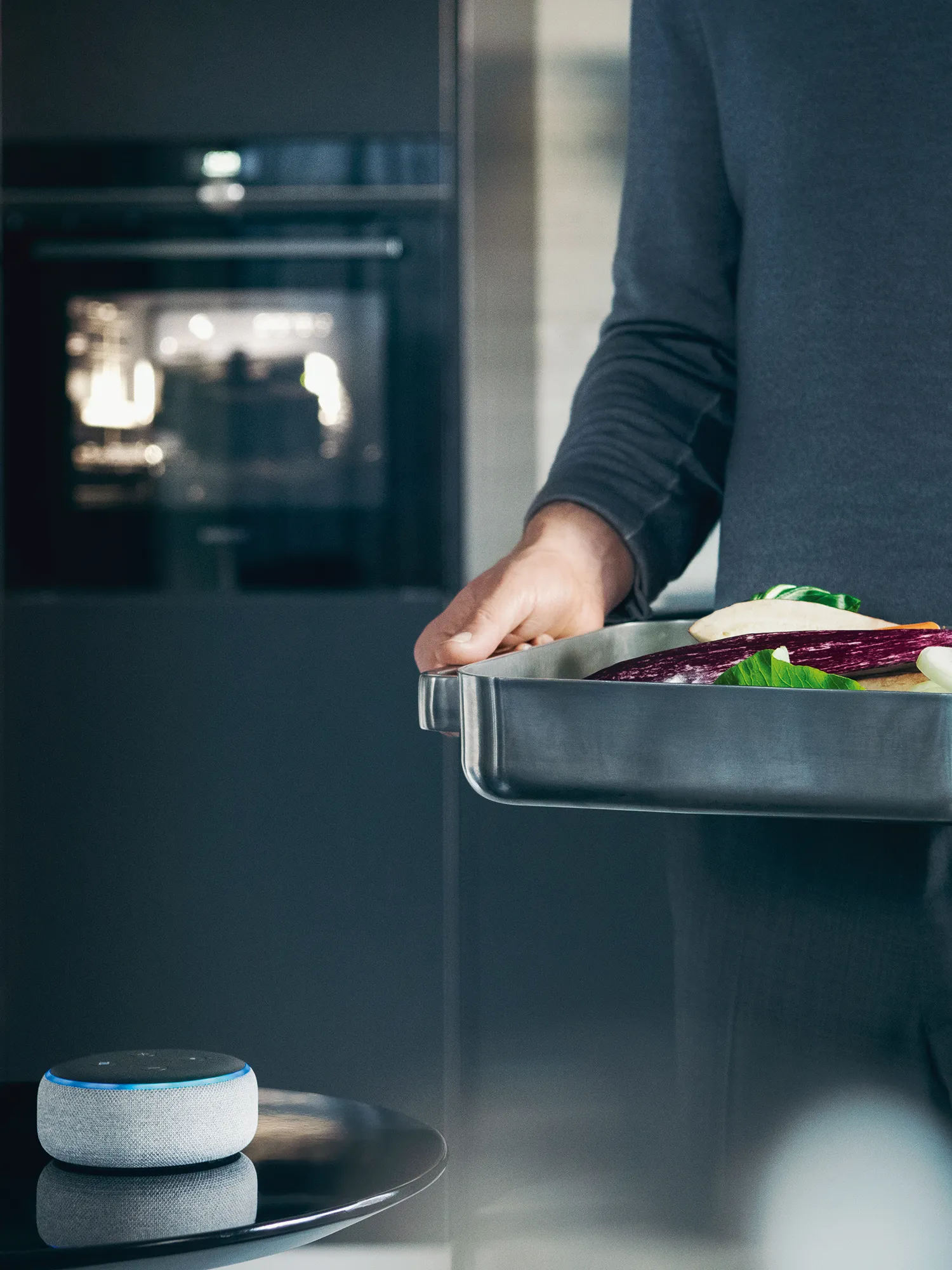Person placing food tray in oven, smart speaker on counter.