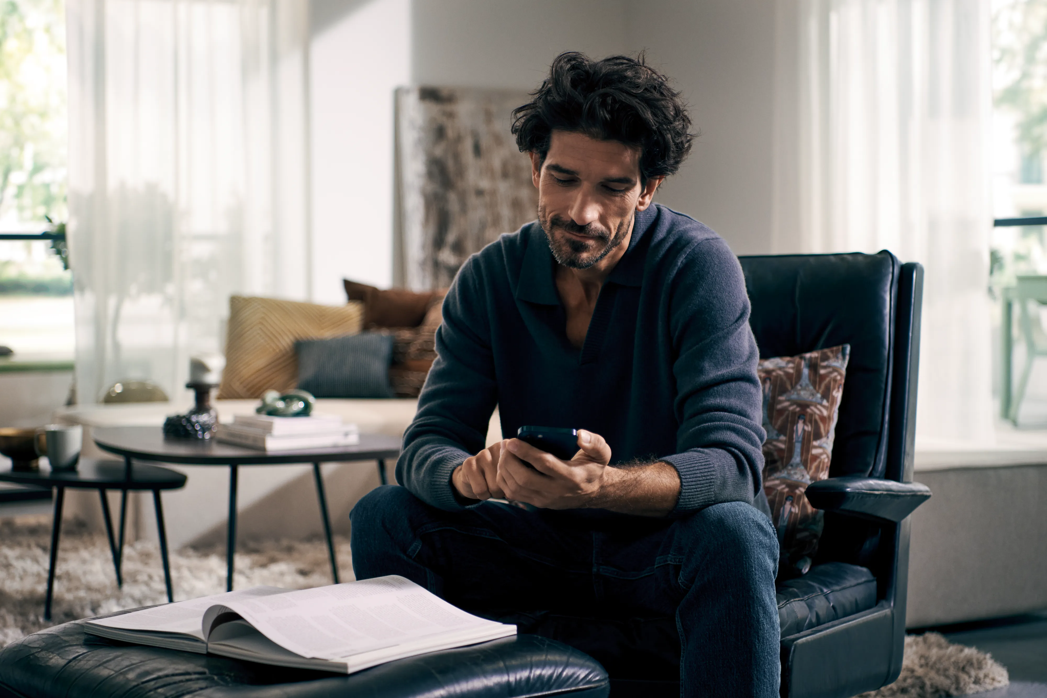 En man sitter i en fåtölj, fokuserad på sin smartphone, framför honom ligger en tidning