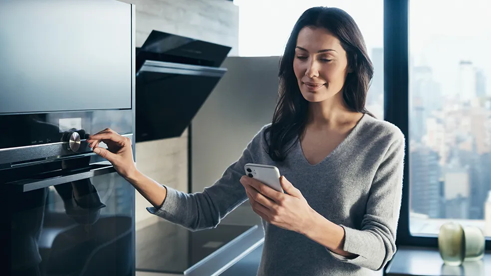 Woman in gray sweater using smartphone while standing in kitchen.