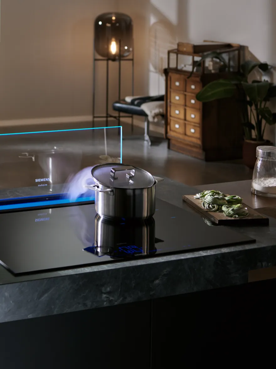 Moderní kuchyně má elegantní indukční varnou desku s hrncem, ze kterého stoupá pára, osvětlenou jemným světlem a s čerstvými surovinami poblíž.